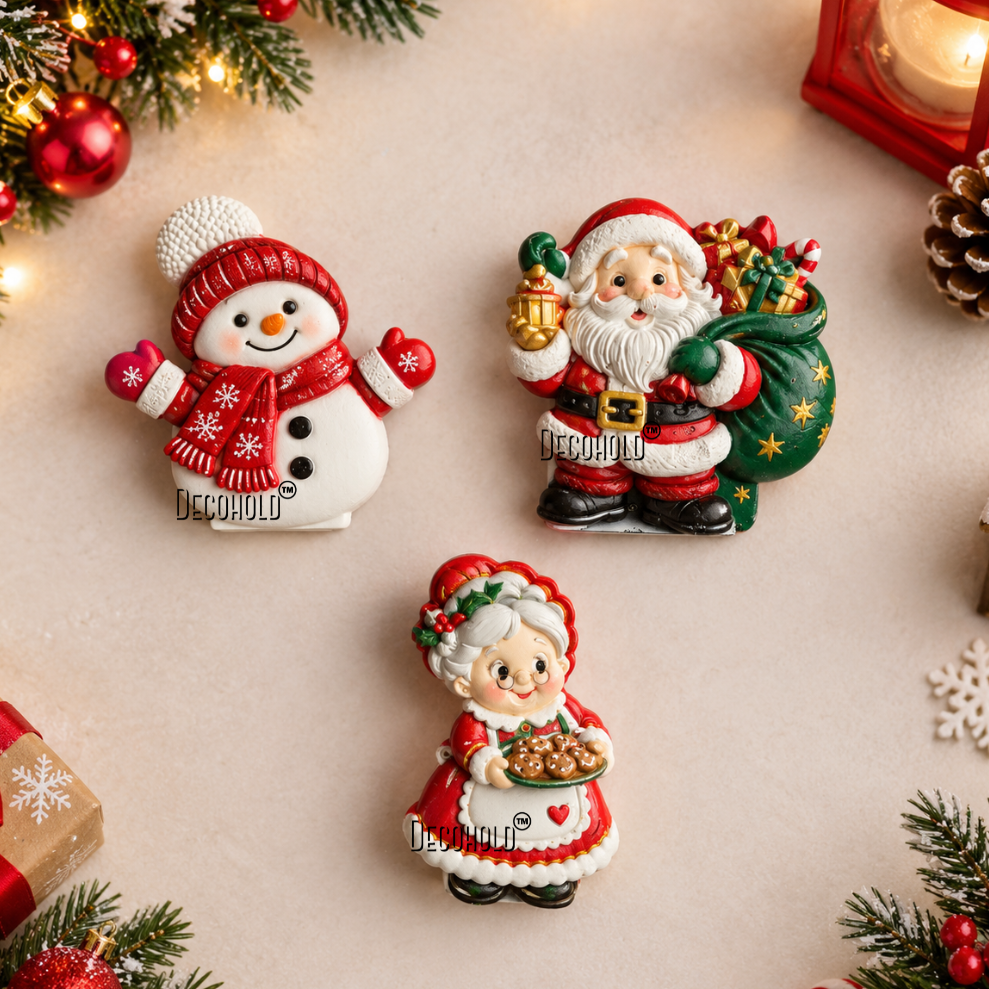 Three Christmas-themed resin figurines displayed on a light beige surface with holiday decorations: a snowman wearing a red hat and scarf, Santa Claus carrying a green sack, and a woman in a red and white dress. The background includes pine branches, ornaments, a lantern, and wrapped gifts.