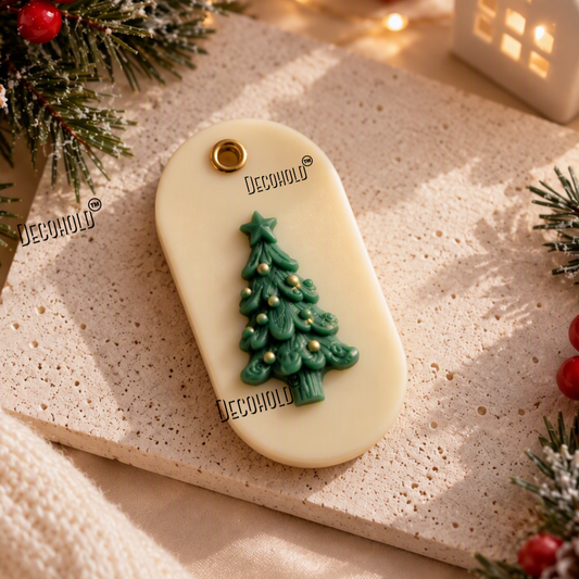 A cream-colored oval pendant or tag with a green Christmas tree charm and small gold accents, styled on a textured beige stone slab with holiday greenery, red berries, and warm string lights in the background.